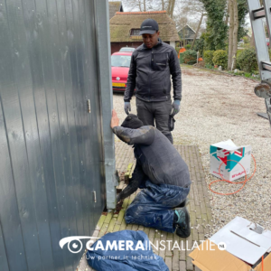 Monteur zit gehurkt bij de onderkant van een houten gevel, met materiaal naast zich op het grind