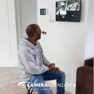 Wandscherm met vier camerabeelden naast elkaar in een woonkamer, met een collega ervoor
