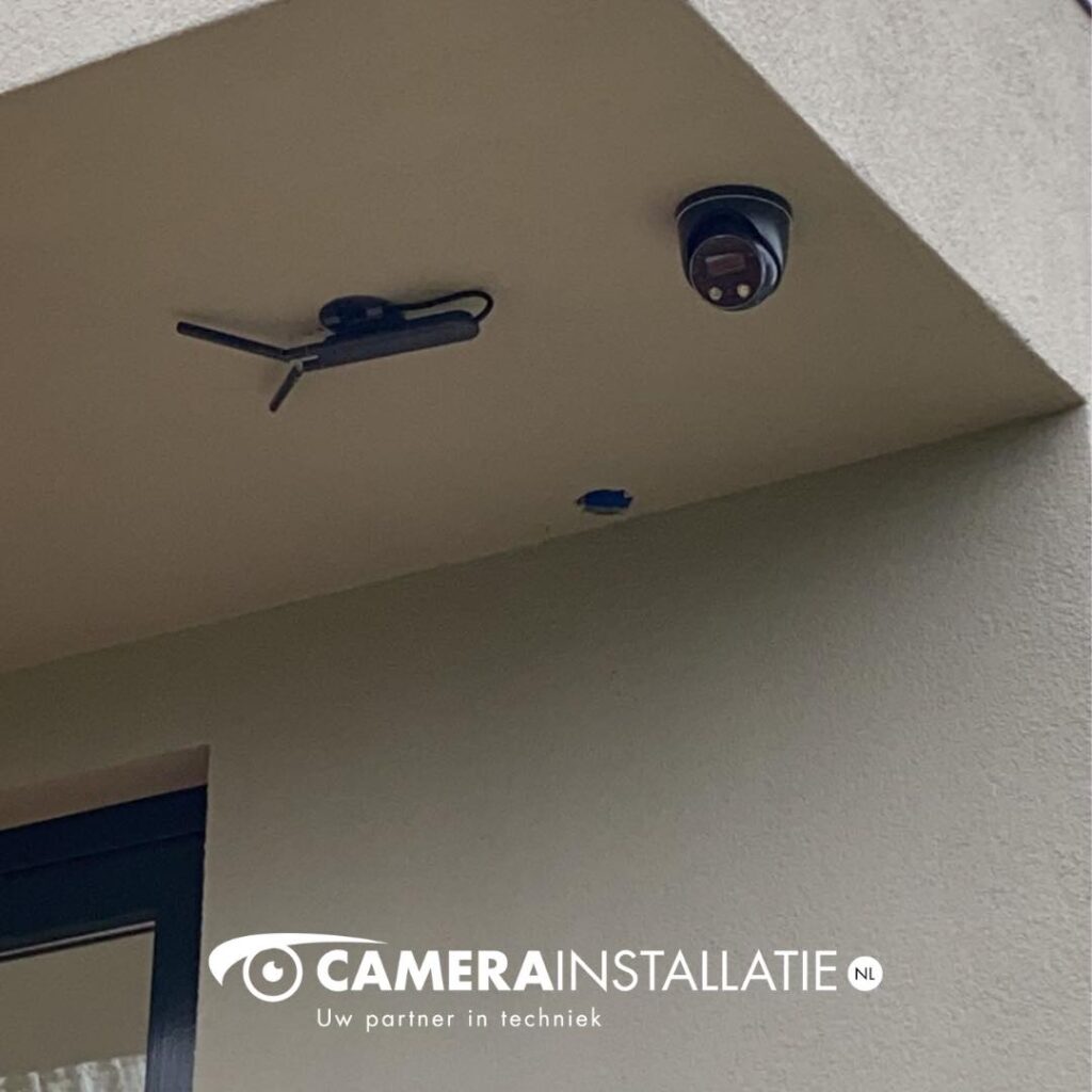 Zwarte turretcamera onder het plafond van het dakoverstek, naast een zwarte buitenspot
