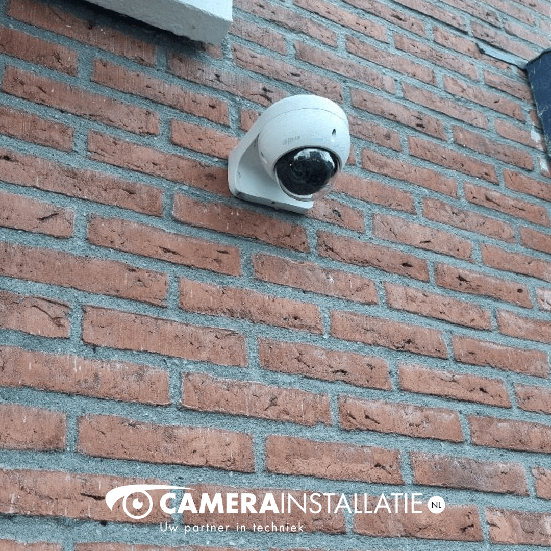 Witte domecamera in een beugel tegen een bruine bakstenen gevel, vlak onder een grijze dakrand