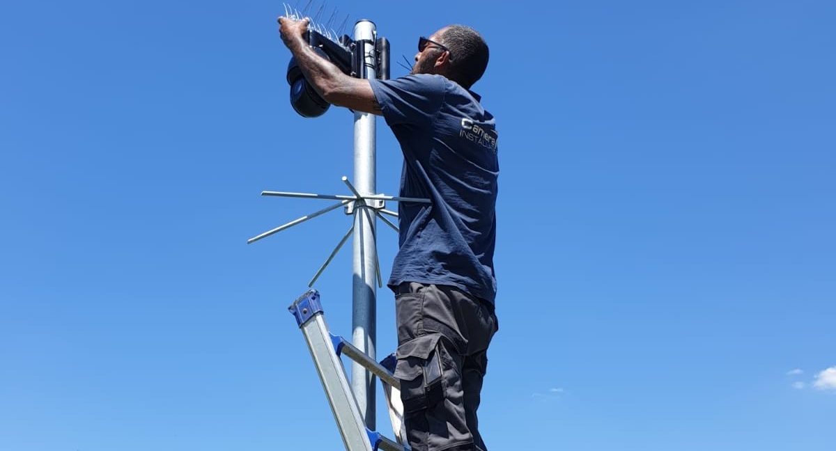 Monteur op een ladder monteert een camera bovenop een mast met duivenpinnen, tegen een blauwe lucht