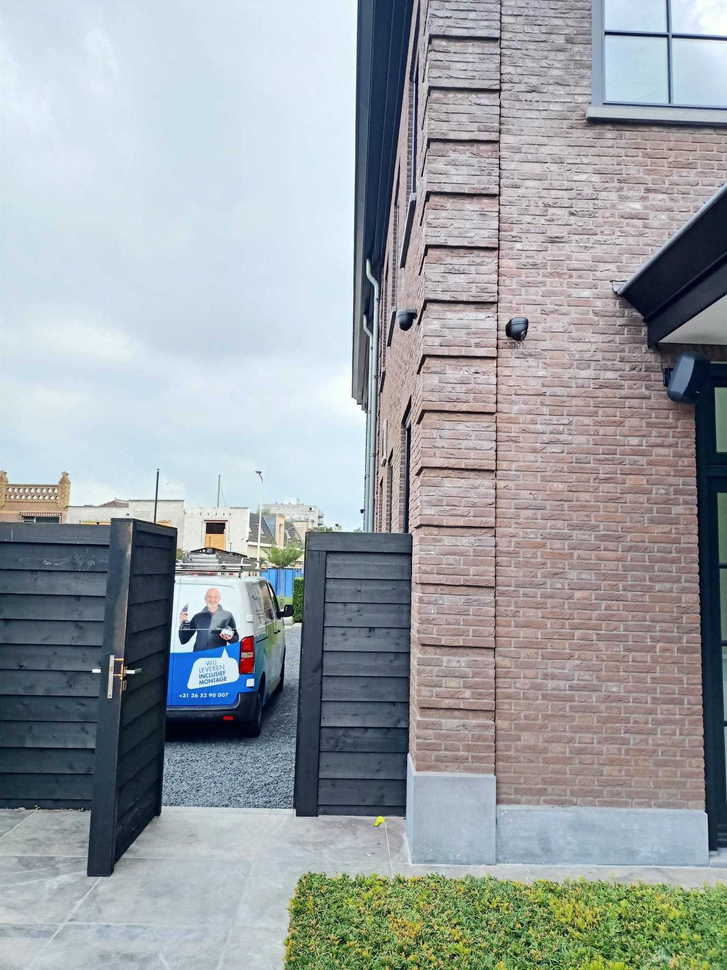 Geopend houten hek naast een bakstenen woning, met hoog op de zijgevel een donkere dome camera en onze bus op de oprit