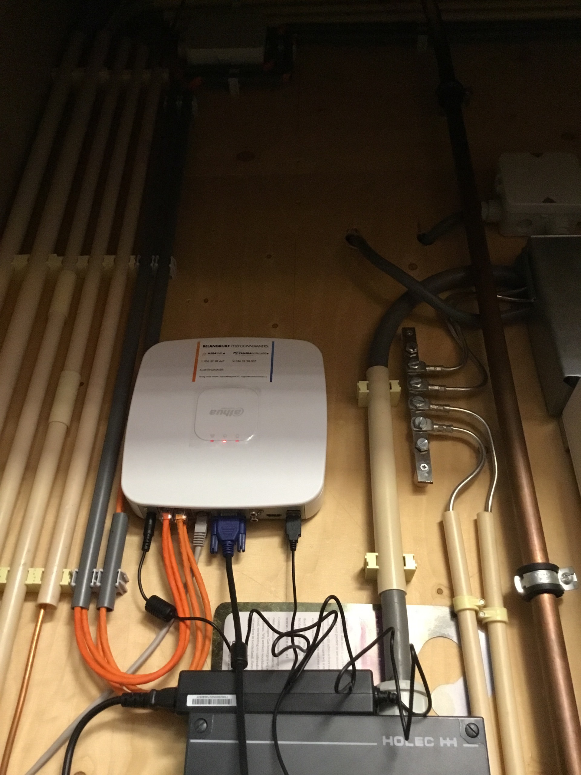 Witte netwerkrecorder op een houten montageplaat in een technische kast, met oranje netwerkkabels eronder