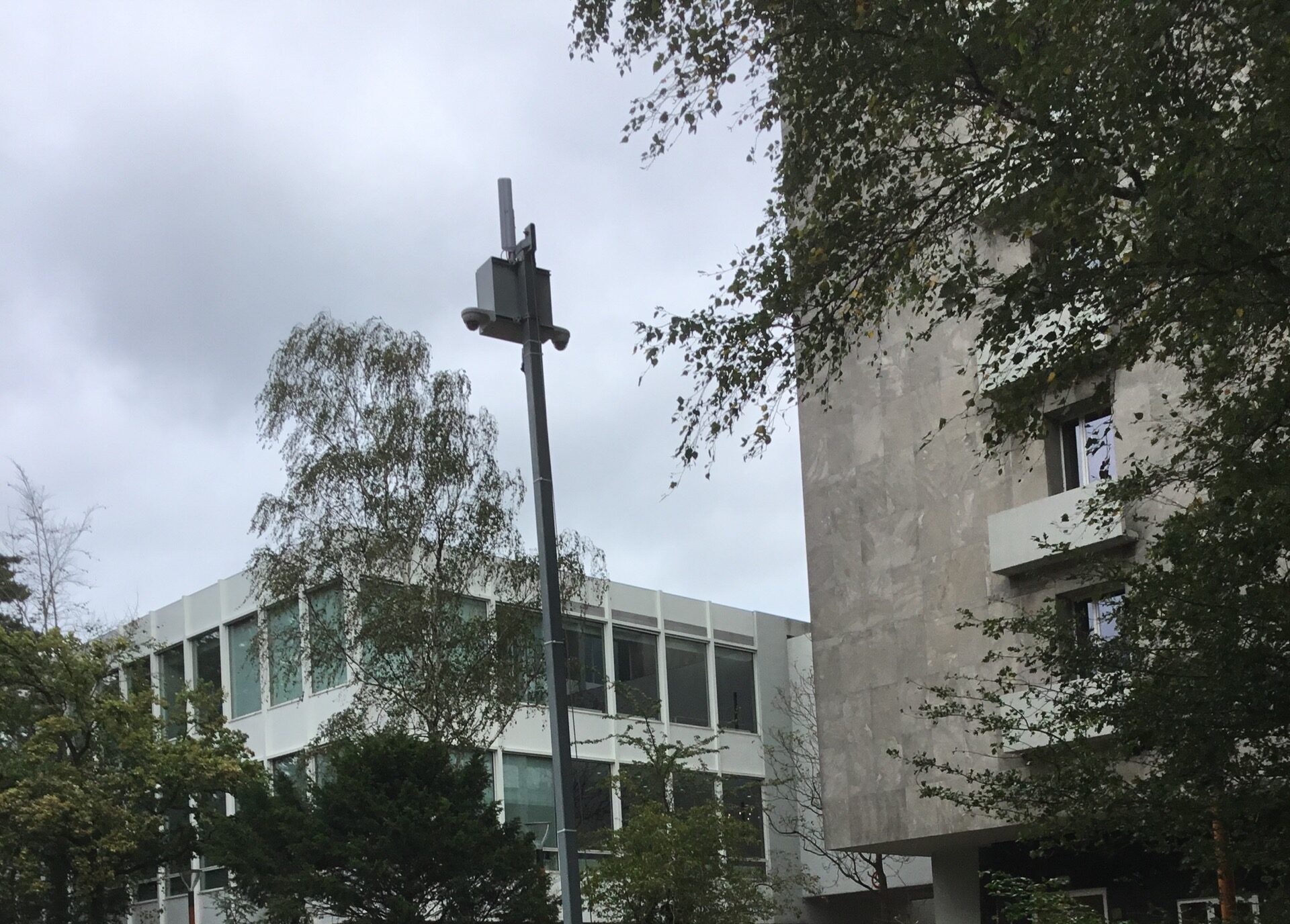 Camera met antennekast bovenop een mast op een schoolterrein, met het gebouw en bomen op de achtergrond