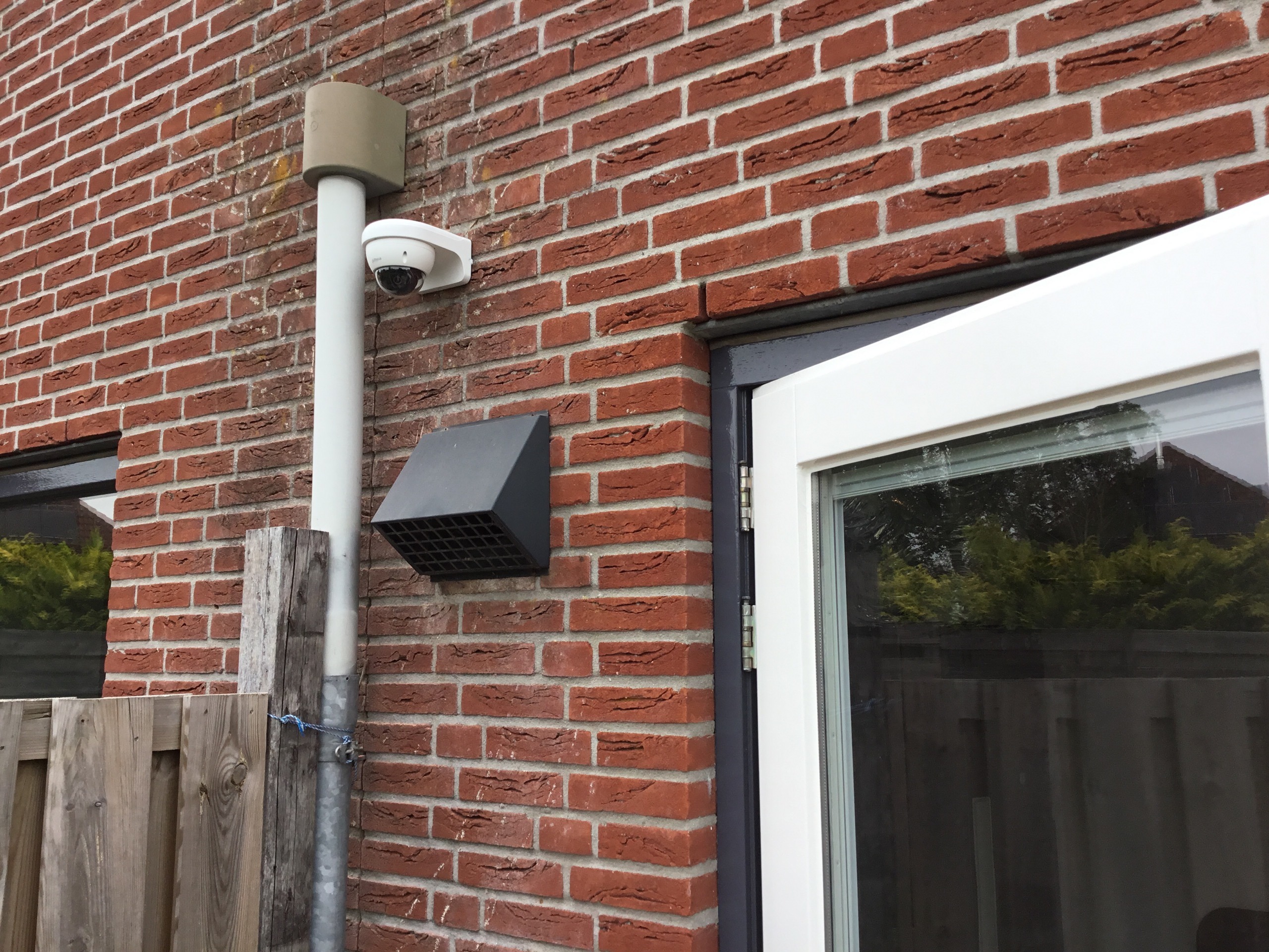 Witte camera tegen een staande buis op een rode bakstenen achtergevel, naast een ventilatierooster en de tuinpoort