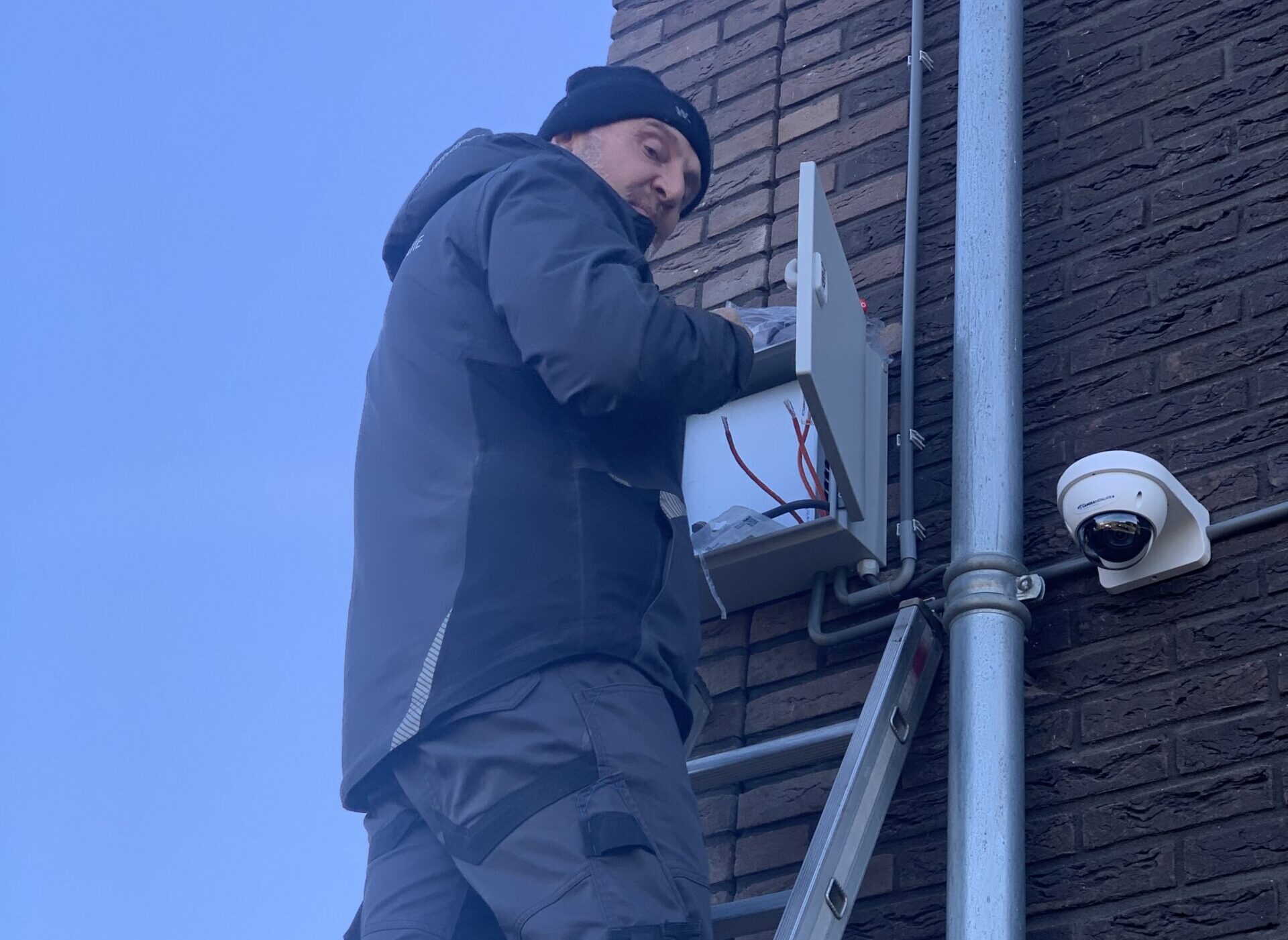 Monteur op een ladder werkt aan een kast naast een camera tegen een donkere bakstenen gevel