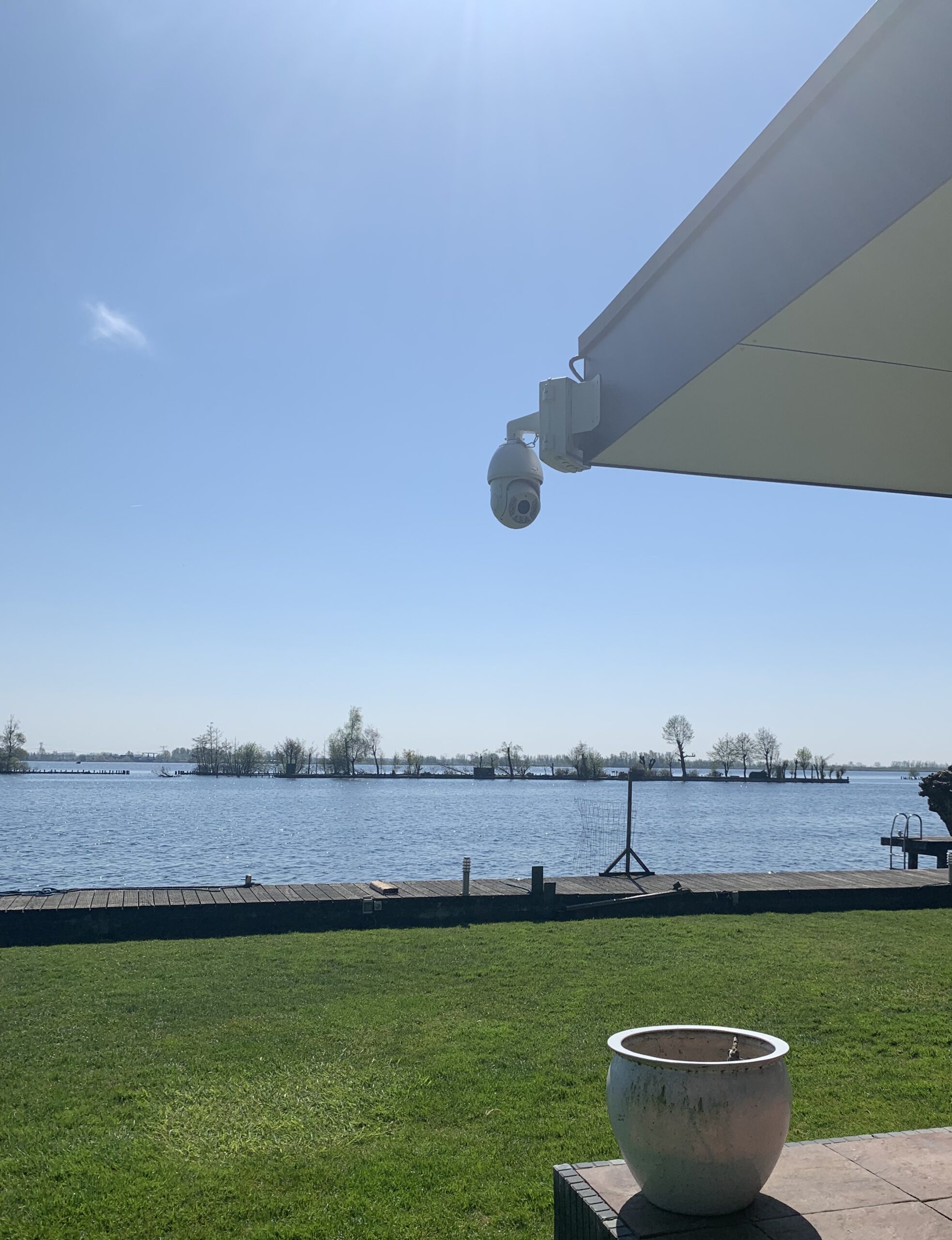 Witte bestuurbare domecamera aan een beugel onder een zonnescherm, met zicht over het gazon, de steiger en het water