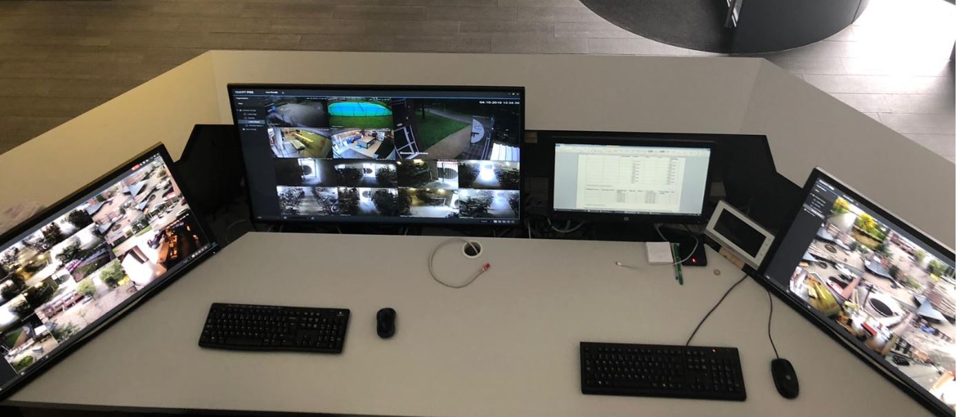 Werkplek in een meldkamer met brede schermen vol camerabeelden, toetsenborden en een klein bedienpaneel
