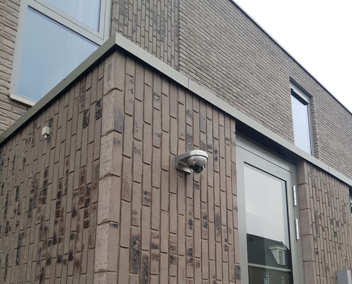 Witte domecamera hoog op de buitenhoek van een moderne bruine baksteengevel naast de voordeur