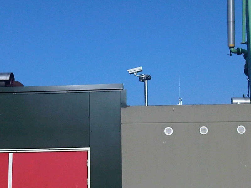 Bestuurbare zoomcamera en een tweede camera op een maatwerkmast aan de dakrand van een bedrijfspand, gericht over het terrein tegen een strakblauwe lucht