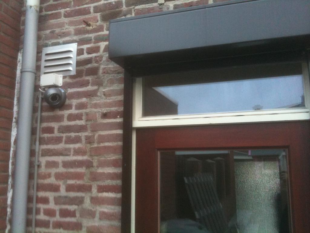 Bewakingscamera gemonteerd op de rode bakstenen gevel van de woning, naast de achterdeur en een regenpijp