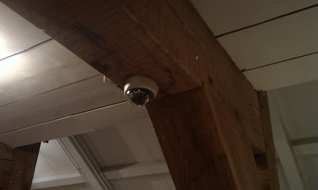 Witte dome-camera gemonteerd tegen een donkere houten balk onder het witte plafond van een boerderij