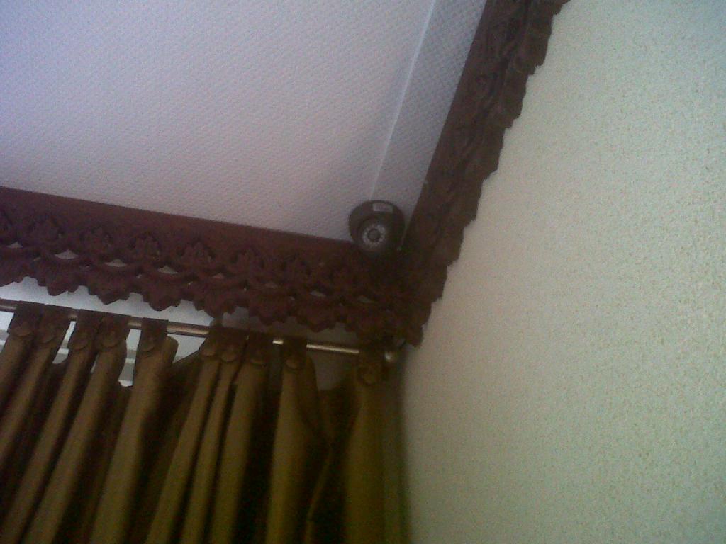 Kleine bewakingscamera discreet in de hoek van een klassieke woonkamer, naast een sierlijke houten gordijnkap
