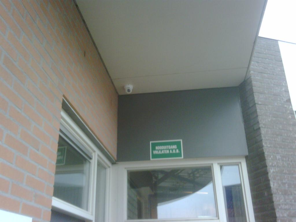 Witte dome-camera onder de dakoverstek op de hoek van een bakstenen pand, boven de ramen bij de entree met een groen nooduitgang-bordje