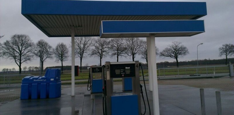 Tankstation met blauwe luifel boven twee brandstofpompen en een nat voorterrein onder een grijze lucht