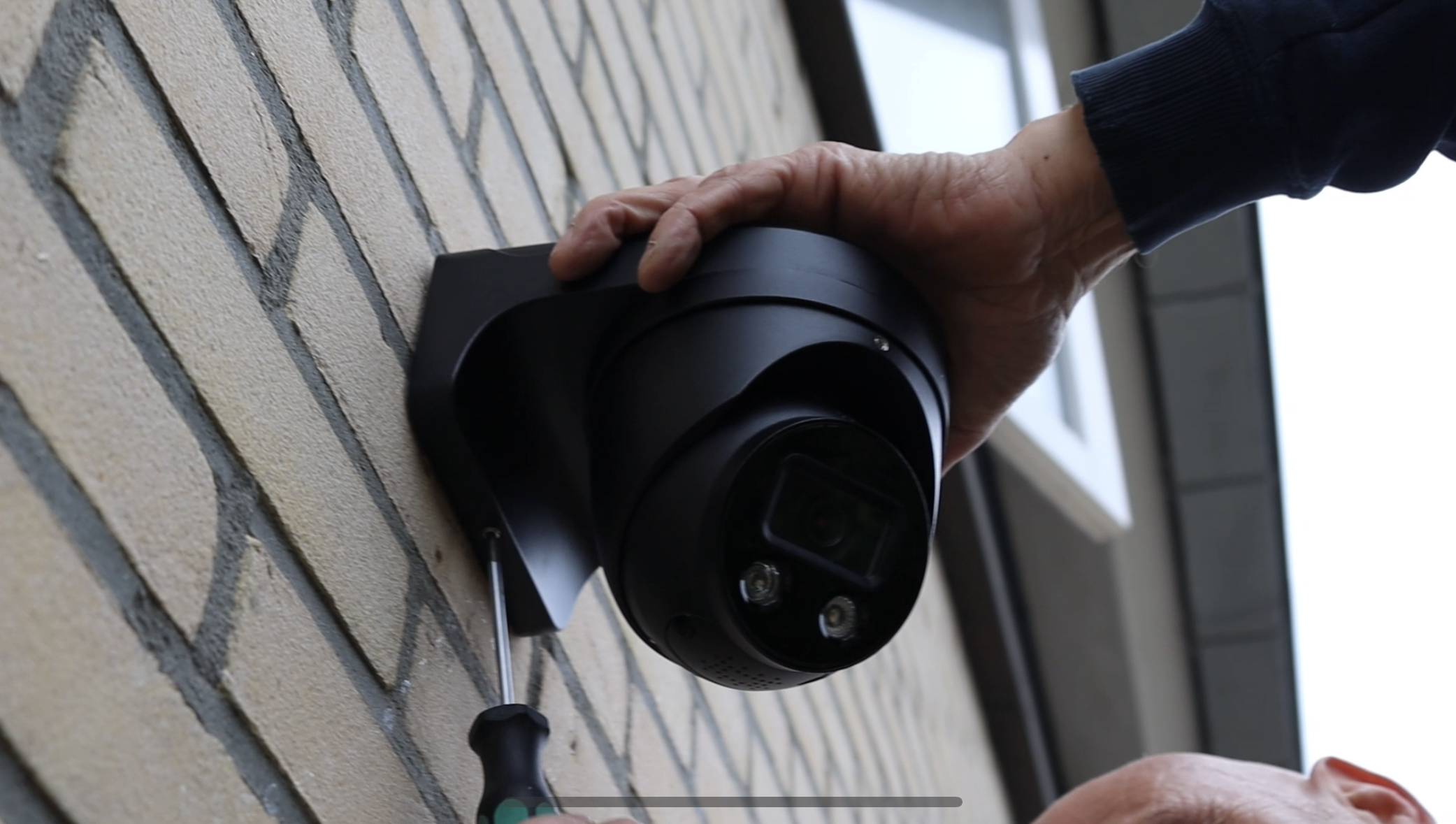 Twee handen zetten met een schroevendraaier een zwarte camera met zonnekap vast tegen een bakstenen gevel