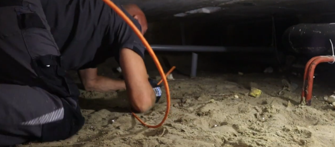 Monteur kruipt met een oranje kabel door een zanderige kruipruimte onder de vloer van een woning
