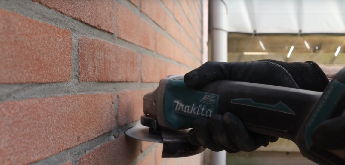Monteur slijpt met een Makita haakse slijper een strakke sleuf in het voegwerk van een bakstenen gevel om de kabel in weg te werken
