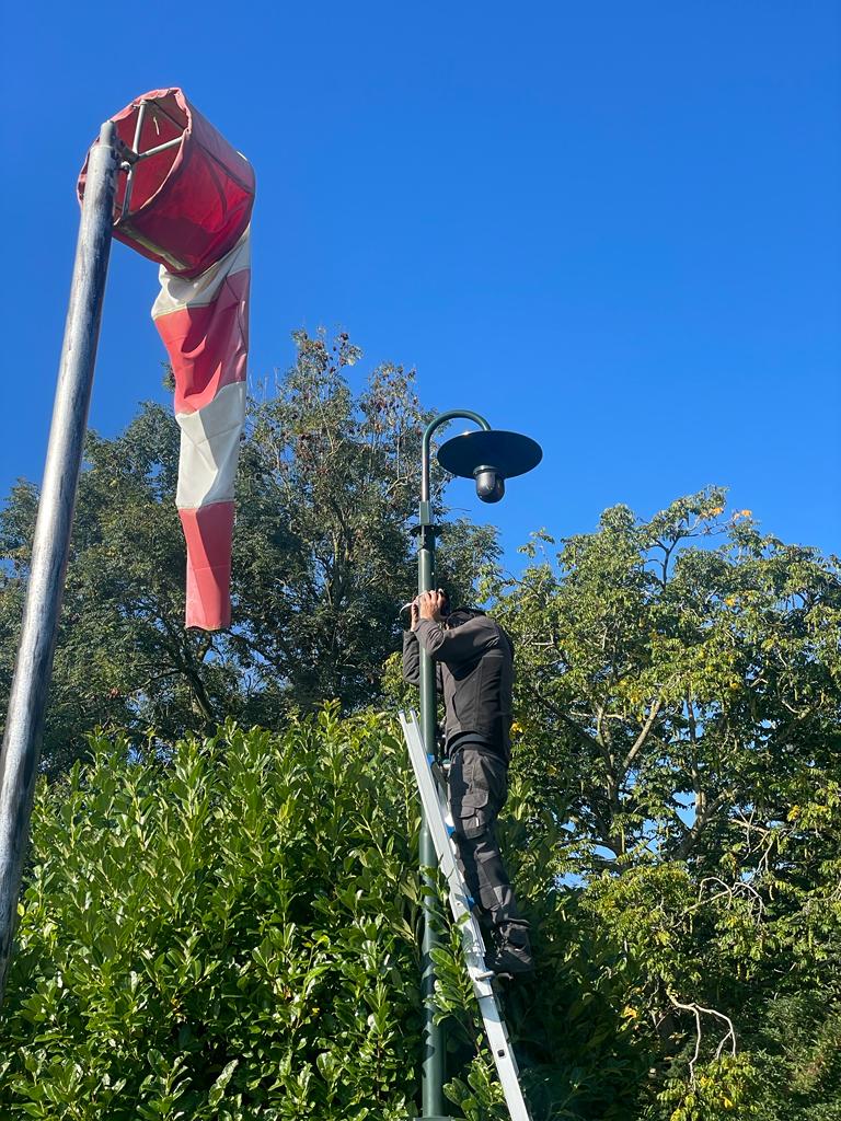 Monteur op een ladder monteert een dome camera onder de kap van een lantaarnpaal, naast een rood-witte windzak