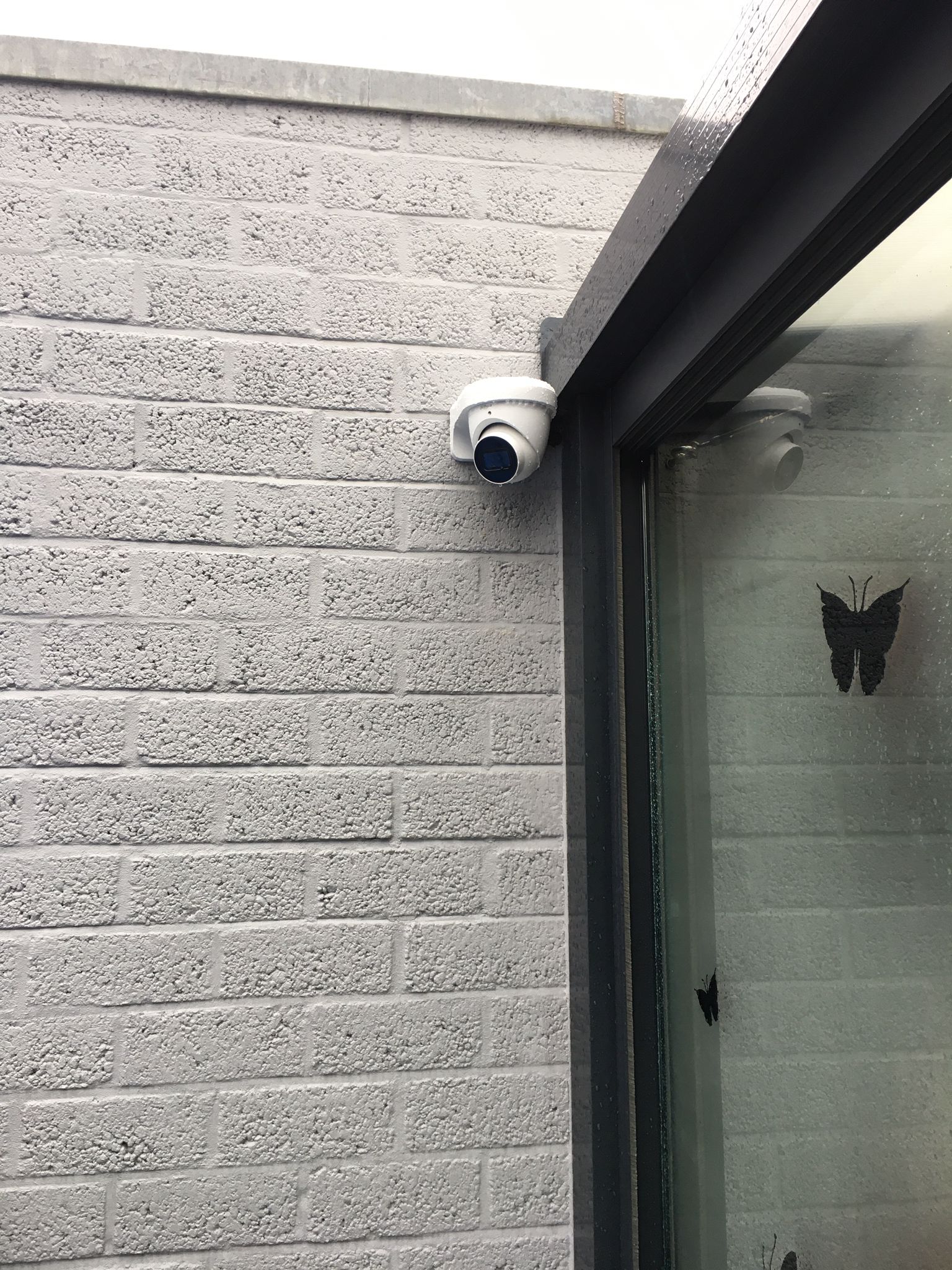 Witte domecamera hoog tegen de lichtgrijze gevel naast het donkere kozijn van de schuifpui