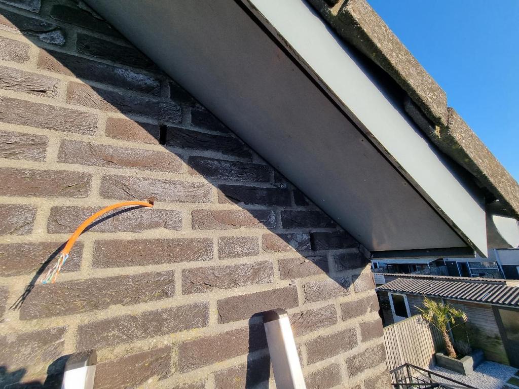Oranje netwerkkabel steekt uit een gat in de voeg hoog op de gevel, vlak onder de dakrand, met een ladder ernaast