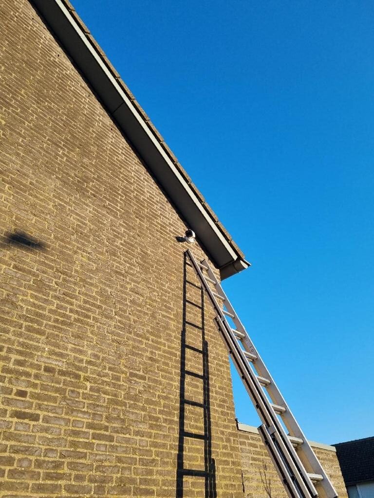 Bewakingscamera hoog op de kopgevel onder de dakrand, met een ladder tegen de gevel en strakblauwe lucht