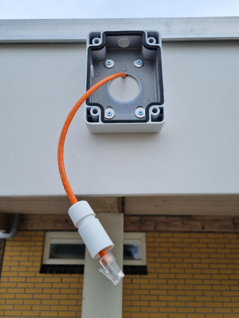 Oranje netwerkkabel met RJ45-connector hangt uit een grijze camera-montagebox onder een dakrand