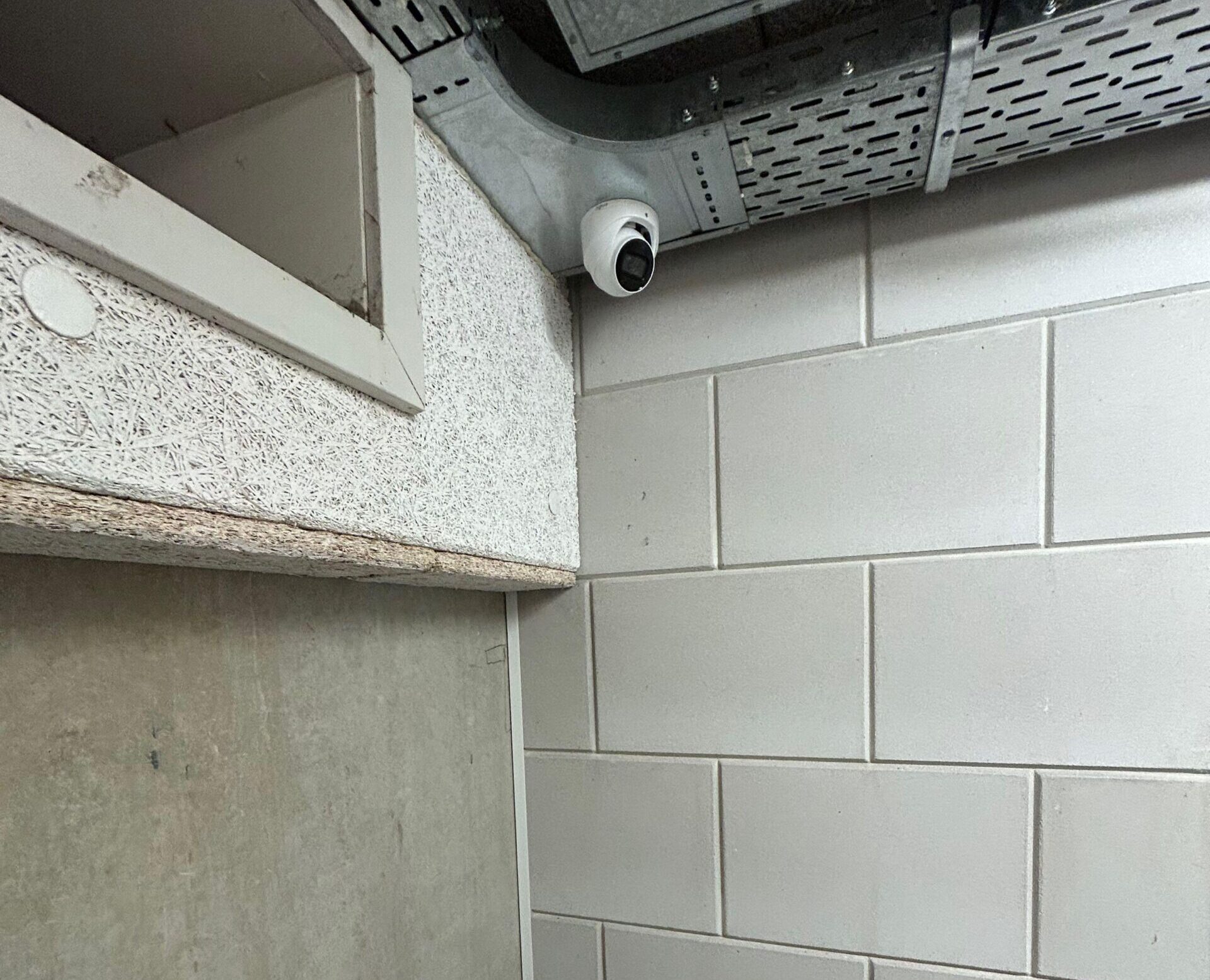 Witte turretcamera net onder het plafond van een bedrijfsruimte, naast een metalen kabelgoot en boven lichte wandtegels