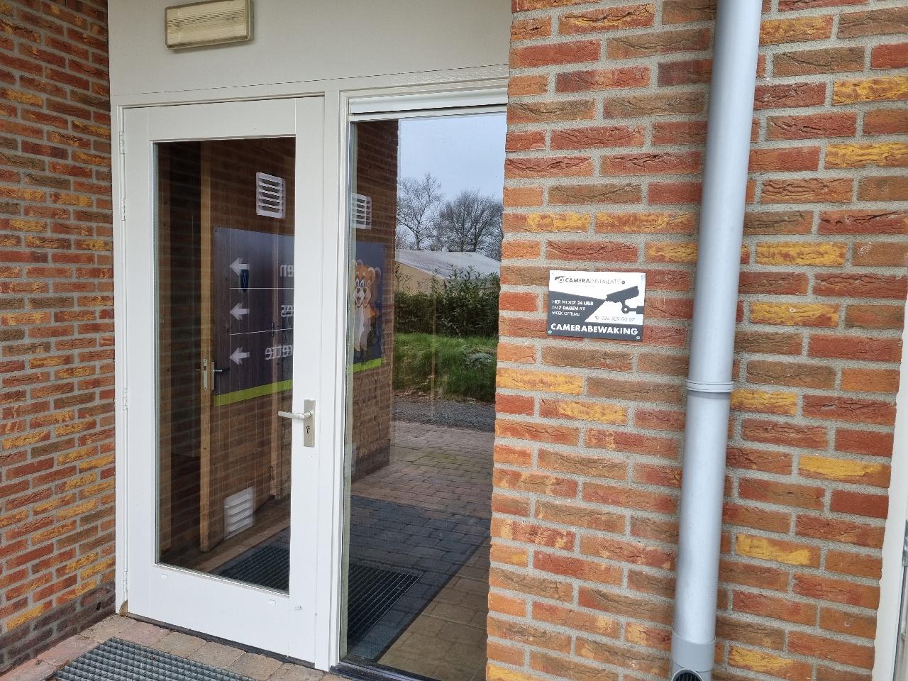 Waarschuwingsbord camerabewaking van CameraInstallatie aan het sanitairgebouw van een camping, met het kampeerveld op de achtergrond