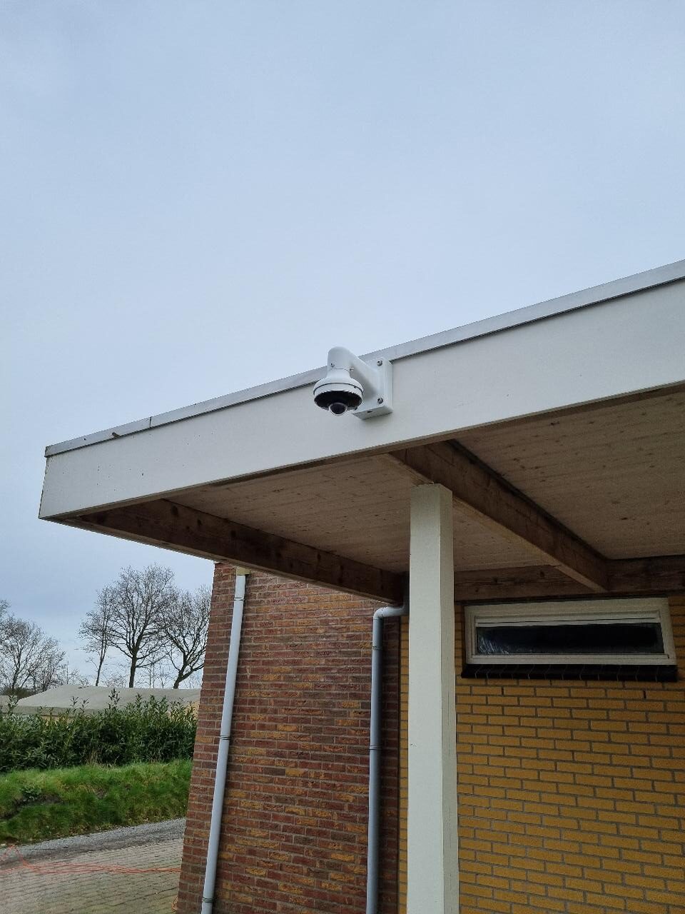 Witte Dahua dome-camera op een wandbeugel onder de dakrand van een sanitairgebouw, gericht op de buitenpaden bij de ingang