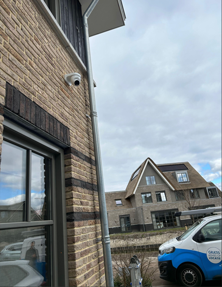 Kleine witte camera hoog aan een bakstenen gevel, met een bedrijfsbus van CameraInstallatie.nl op straat
