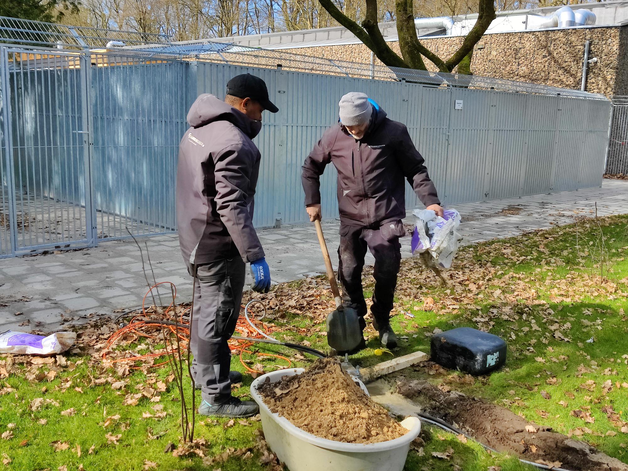 Twee monteurs graven een kabelsleuf in het gras naast de buitenrennen
