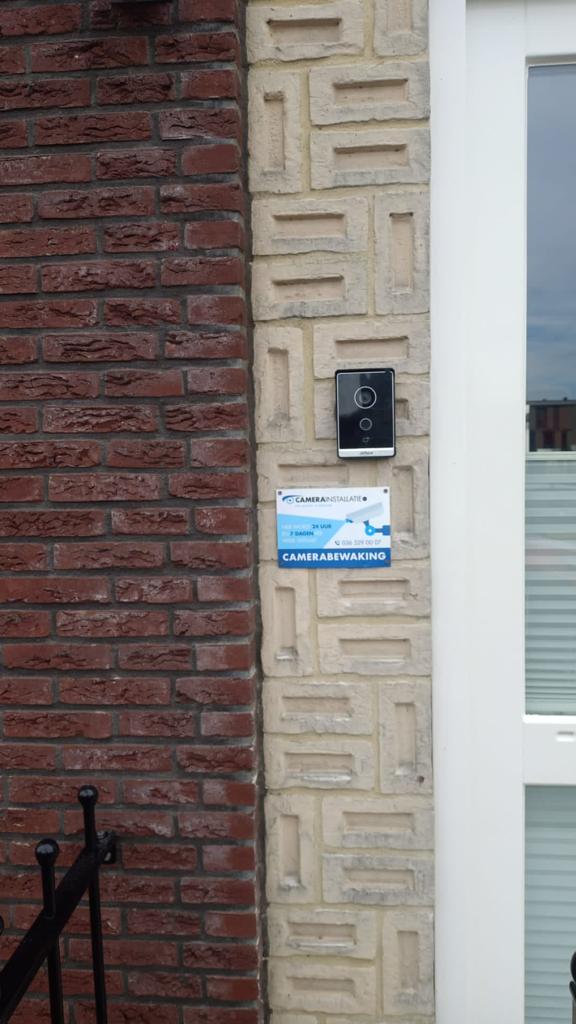 Videodeurbel met camera naast de voordeur van een woning, met een bordje camerabewaking eronder