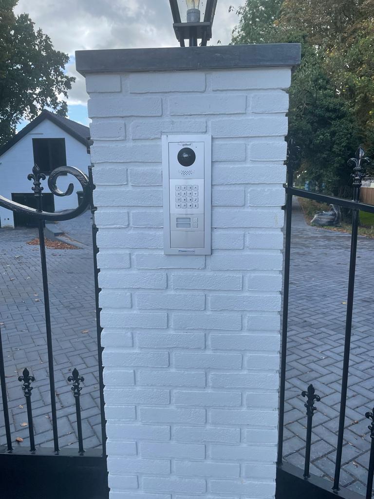 Vervangen intercom met camera en luidspreker, ingebouwd in een witte gemetselde entreezuil bij een gesloten sierhek