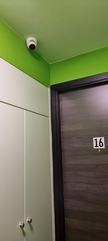 Kleine witte camera in de hoek van een hotelgang bij de lift, boven een kamerdeur met nummer