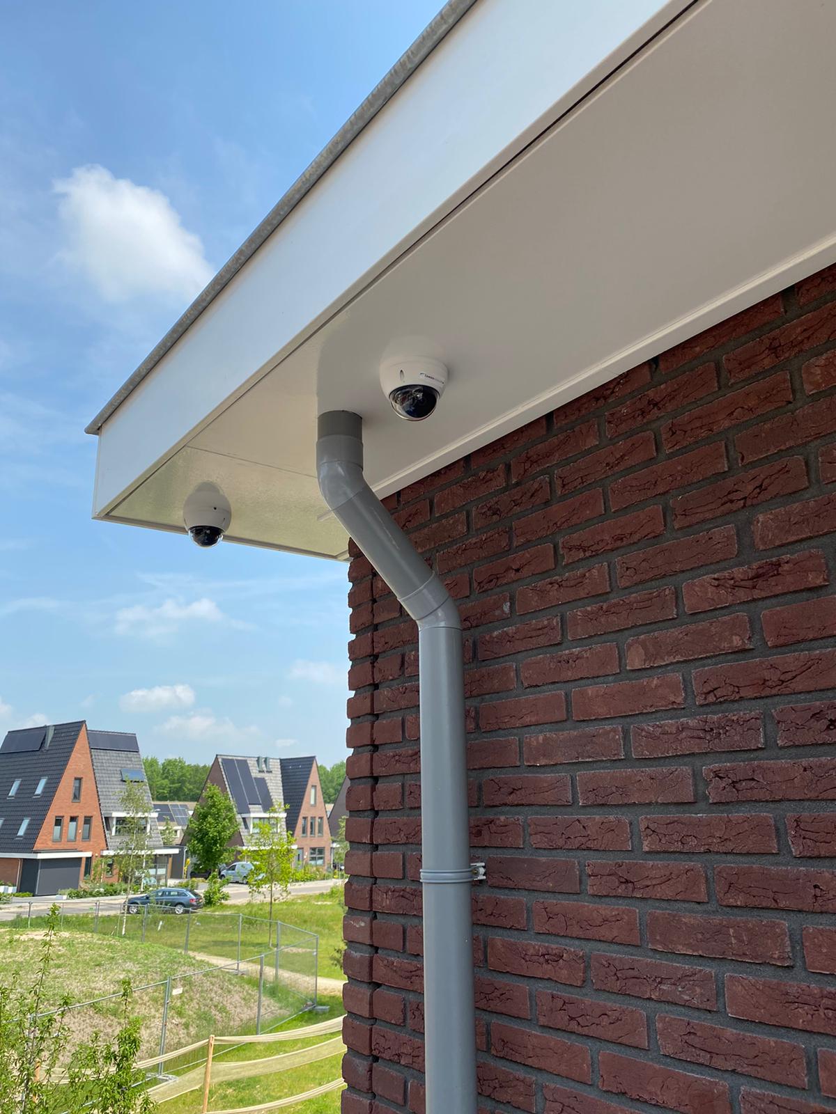 Twee witte dome-camera's onder de witte dakrand van een bakstenen woning, met een nieuwbouwbuurt op de achtergrond