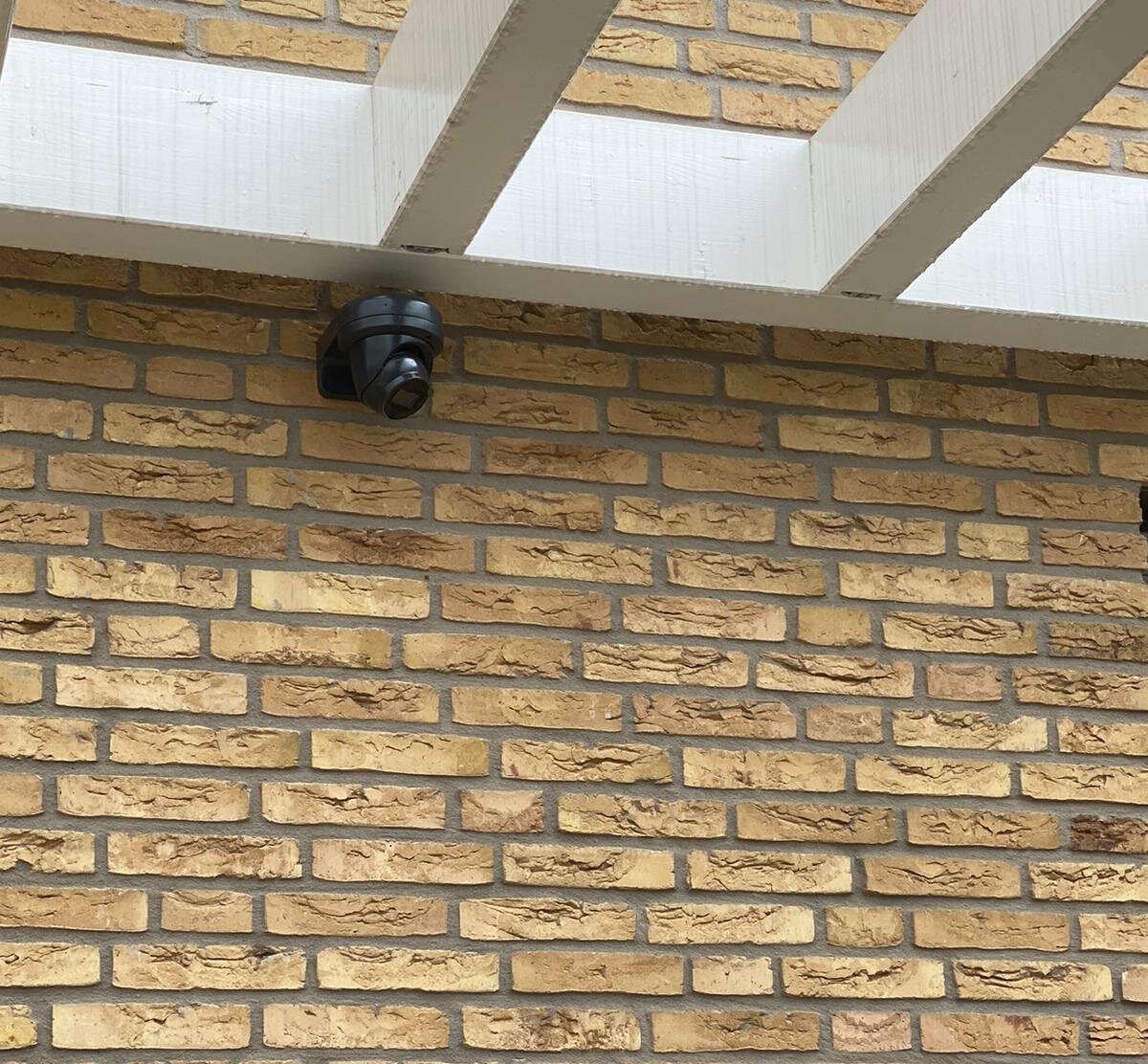 Zwarte camera onder de balken van een overkapping aan een gele bakstenen gevel