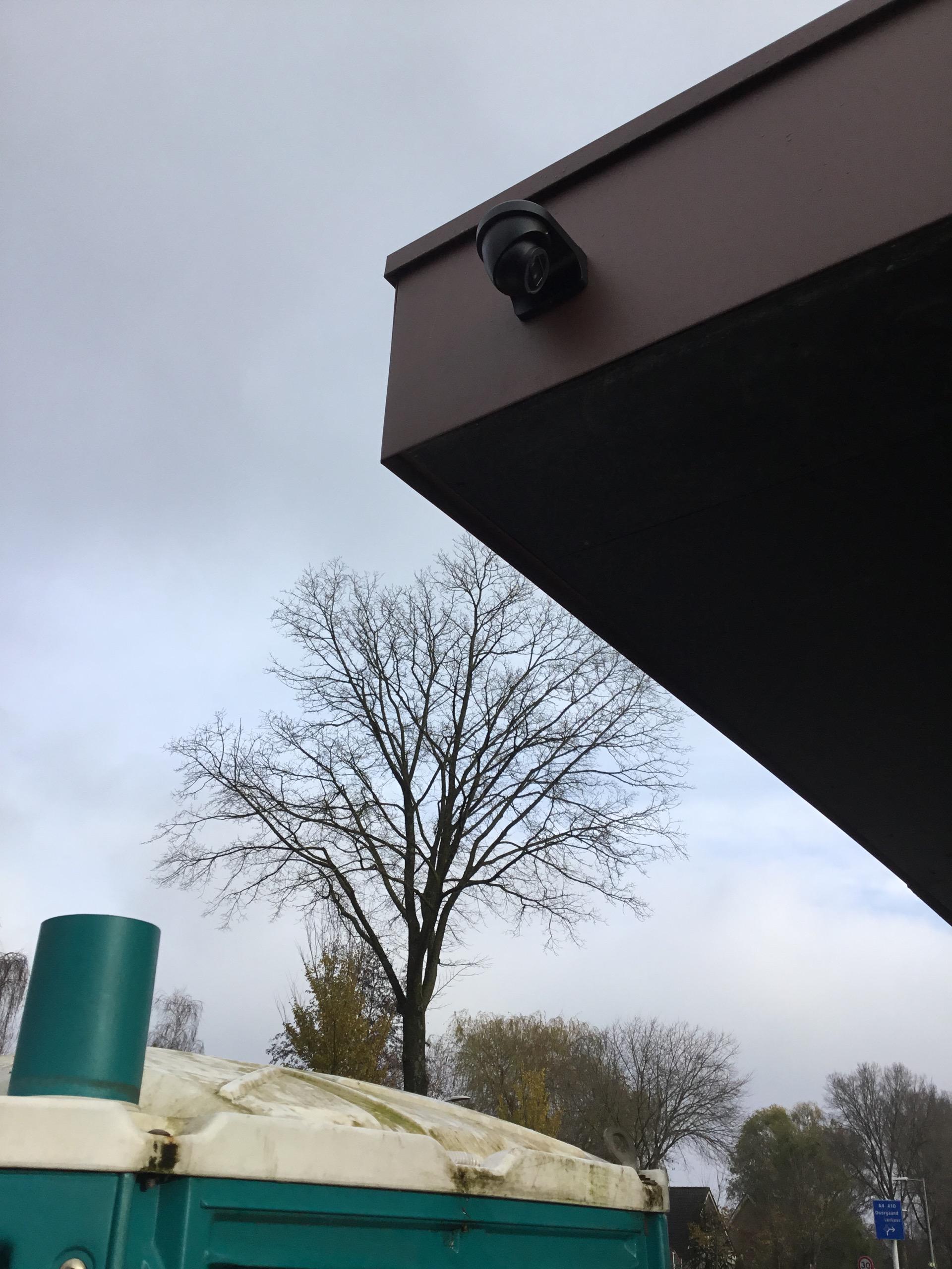 Zwarte dome-camera gemonteerd onder de donkere overstek van een pand, met daarachter een grijze lucht en kale bomen
