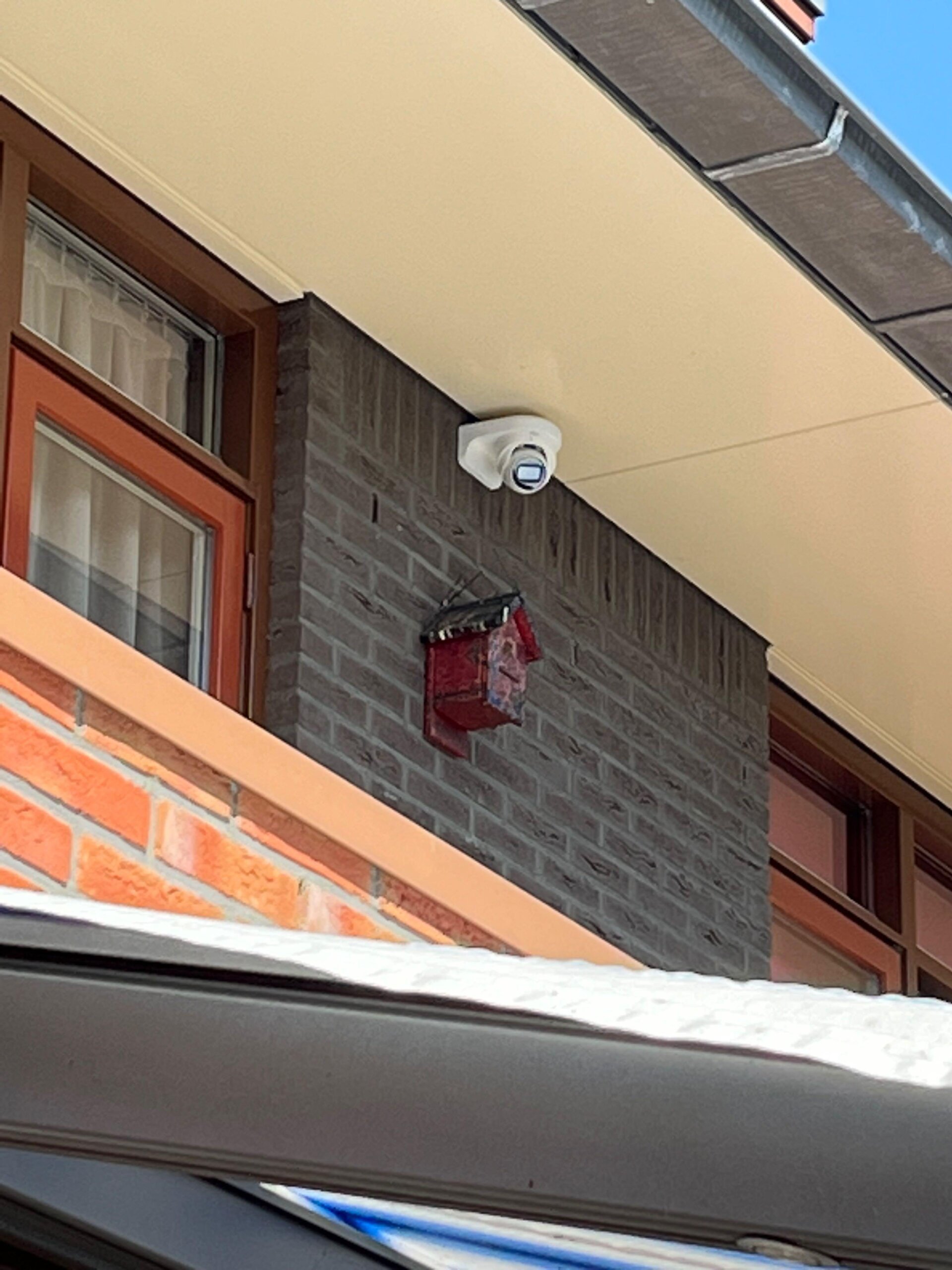 Camera onder de dakrand boven een raam met bruin houten kozijn en een grijze bakstenen strook