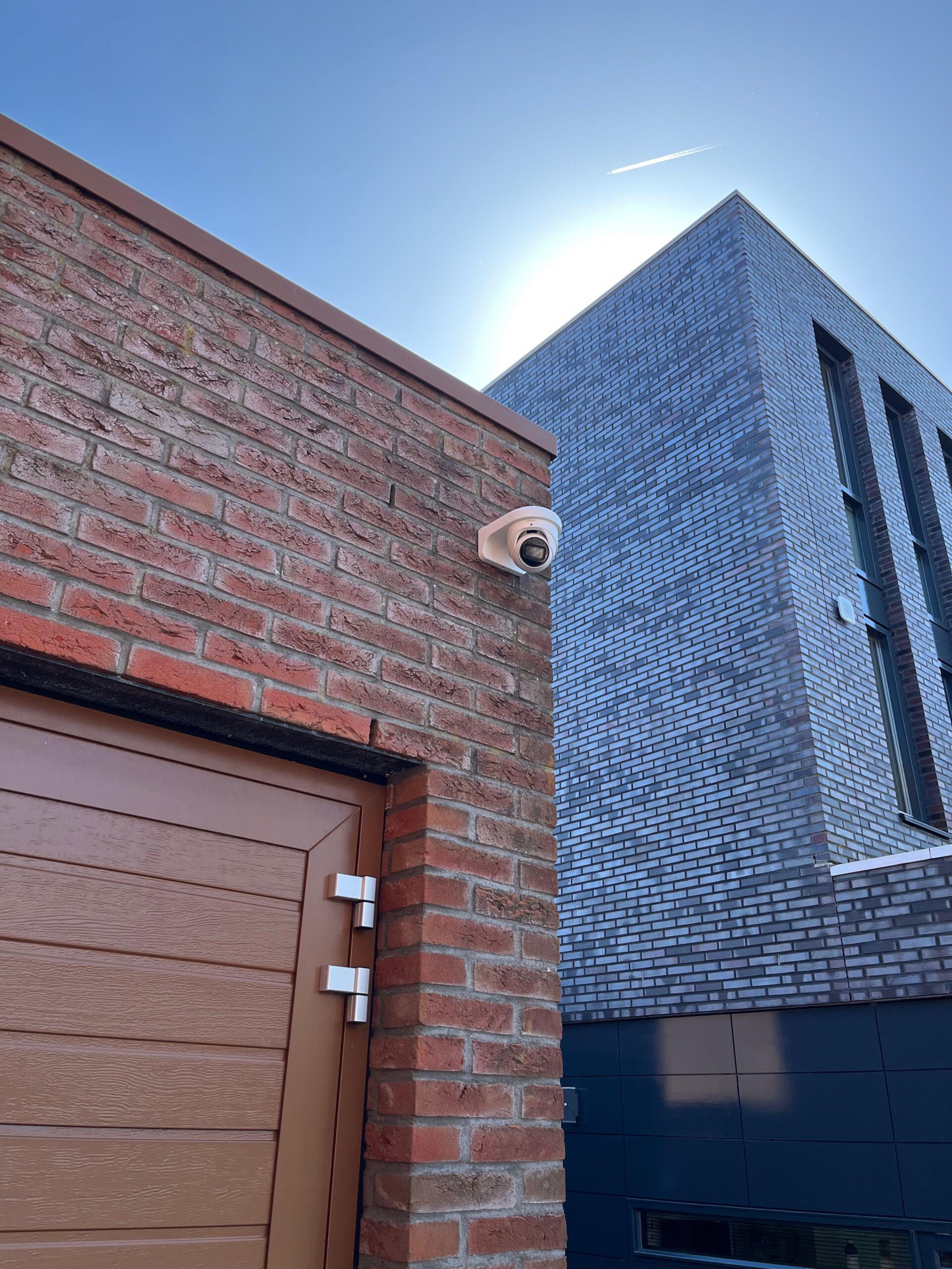 Witte camera op de hoek boven een bruine garagedeur, met strakblauwe lucht en een donkere gevel op de achtergrond