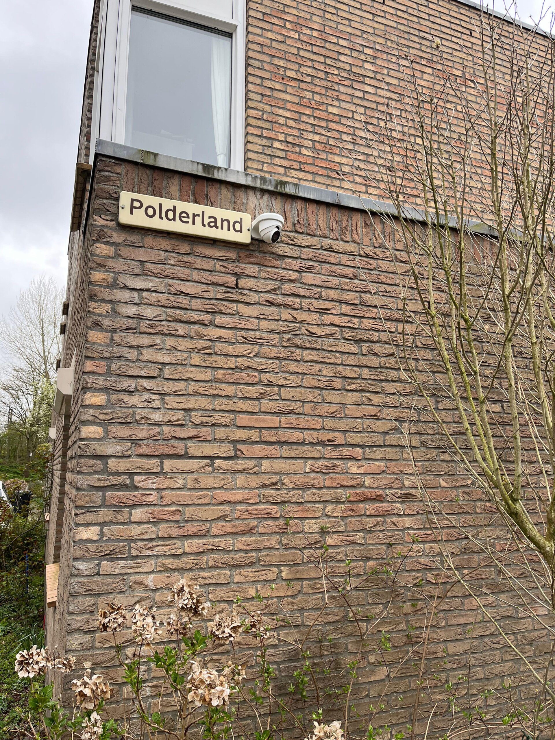 Camera onder een straatnaambord op de blinde kopgevel van een woning, met kale takken ervoor