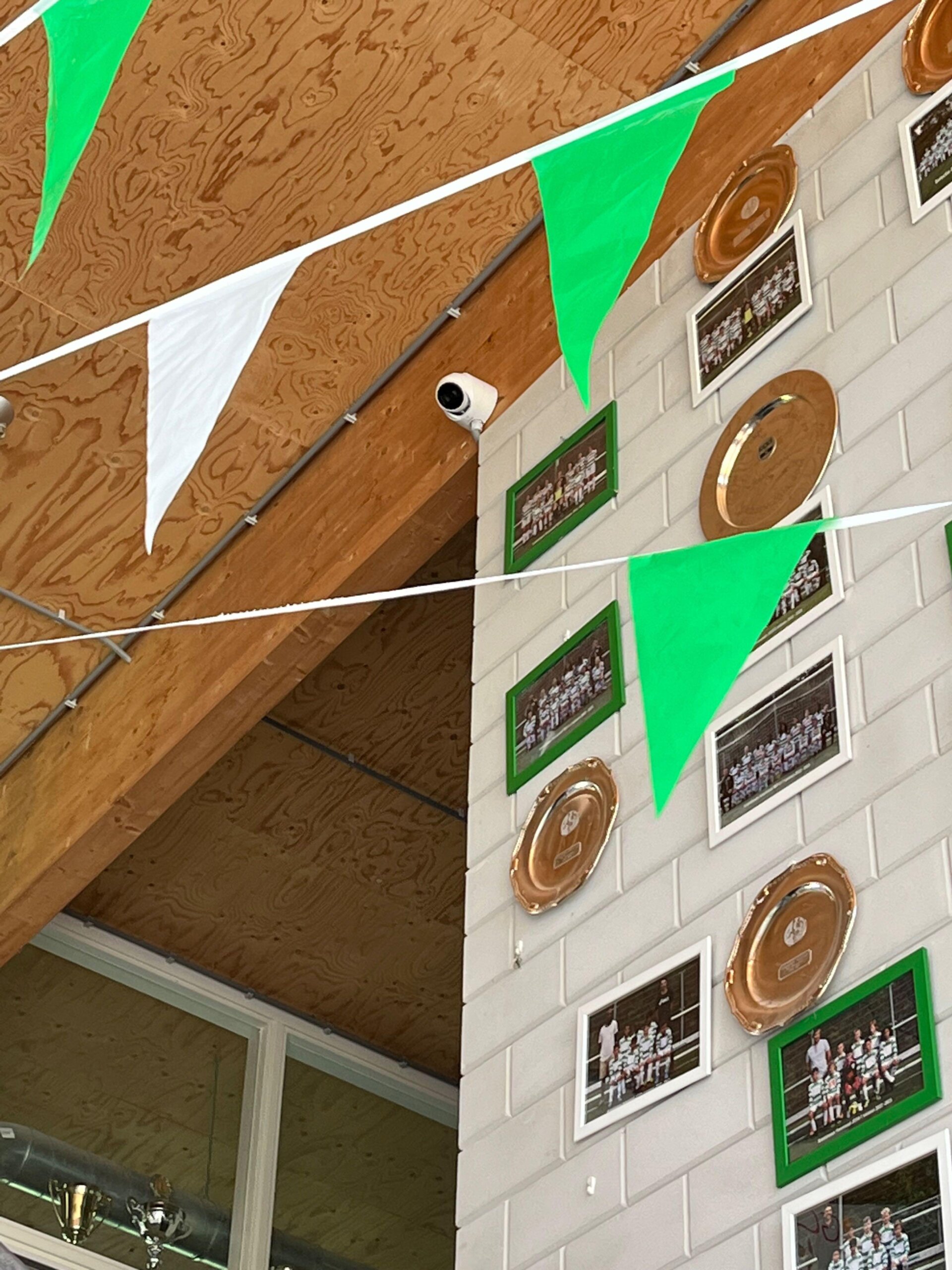 Camera hoog in de kantine, tussen groen-witte vlaggetjes, ingelijste teamfoto’s en gegraveerde schalen