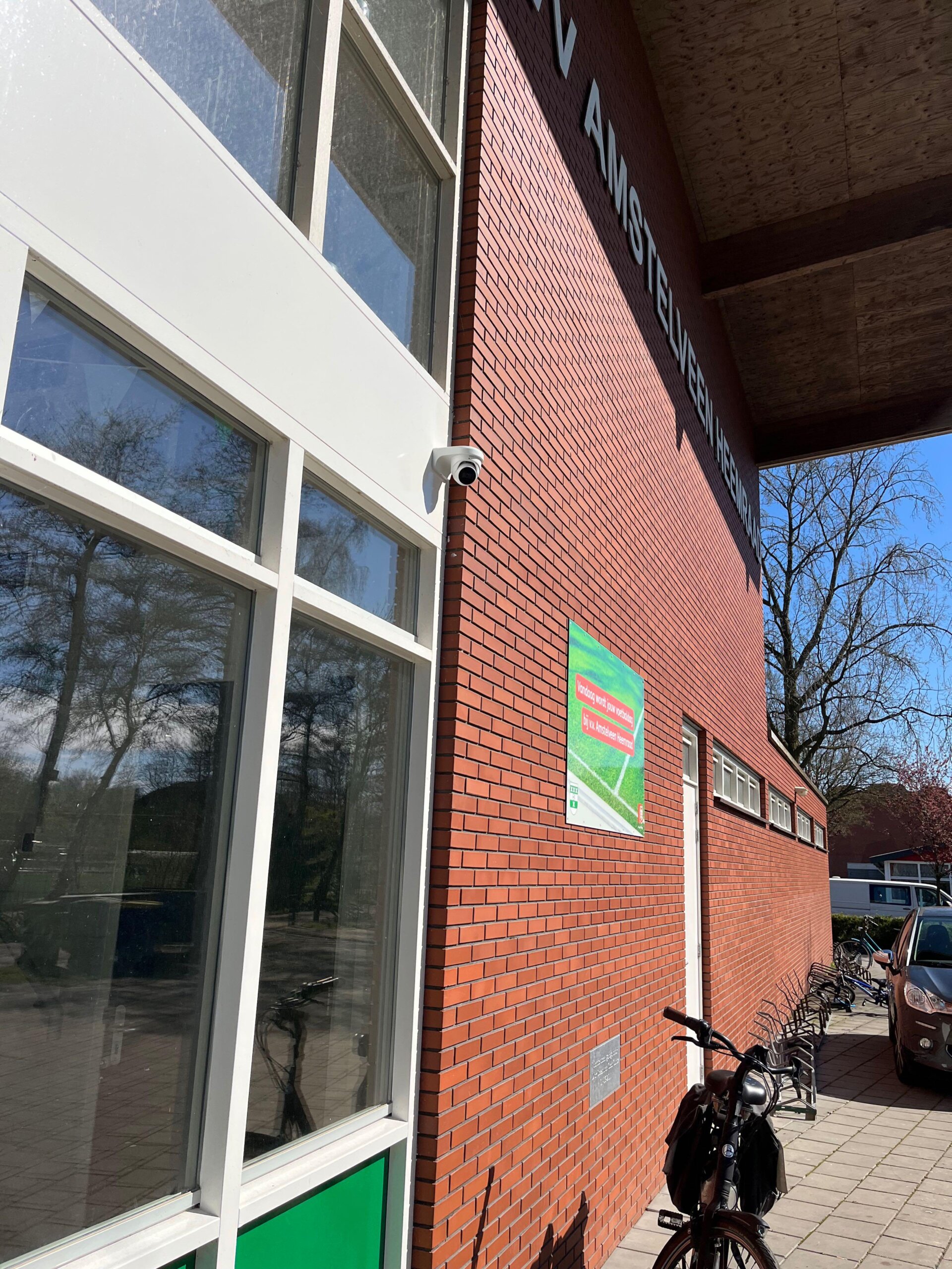 Camera aan de rode bakstenen gevel van het clubgebouw van VV Amstelveen/Heemraad, met fietsenrekken langs het pad