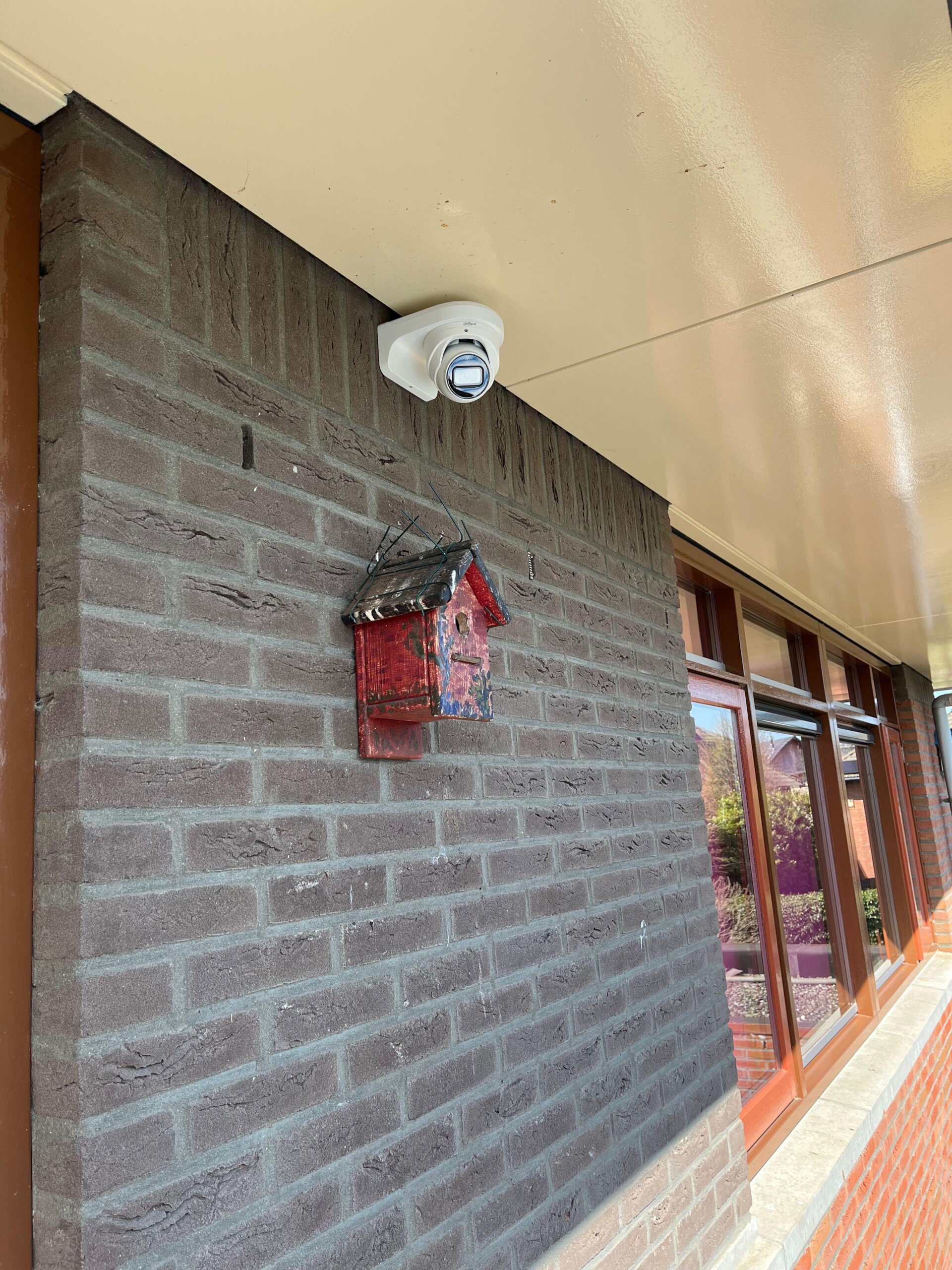Camera onder een lichte plafondafwerking tegen een donkere bakstenen gevel, boven een rood wandobject