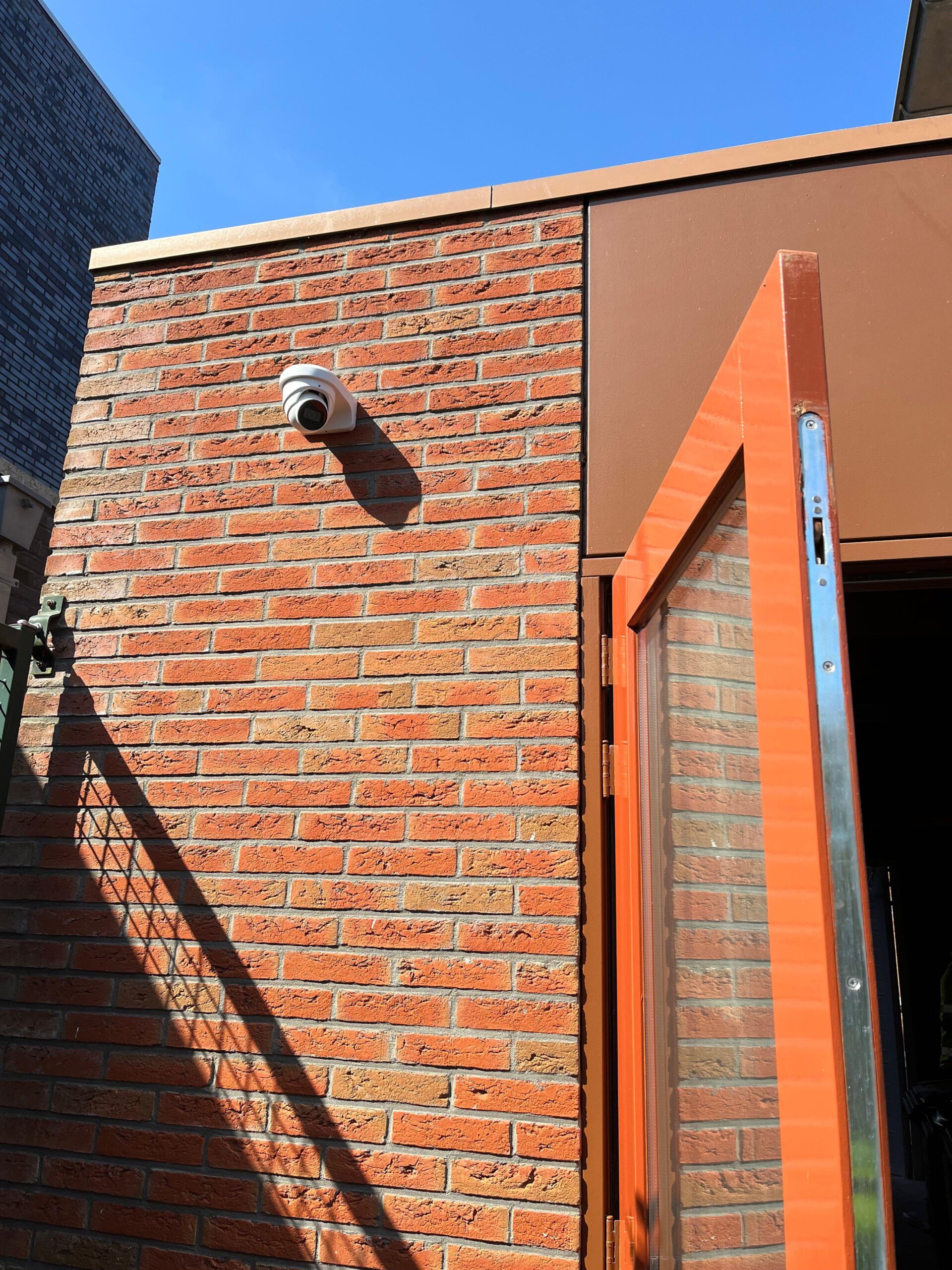 Camera op een oranjerode bakstenen zijgevel naast een openstaande poortdeur, met harde schaduw op de muur
