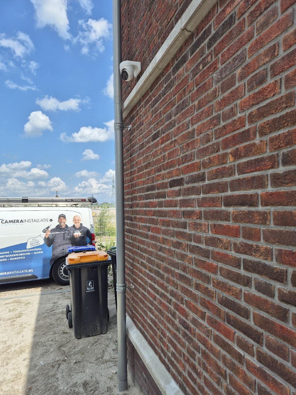 Twee monteurs van CameraInstallatie.nl bij de bedrijfsbus voor een nieuwbouwwoning, met een camera onder de dakrand