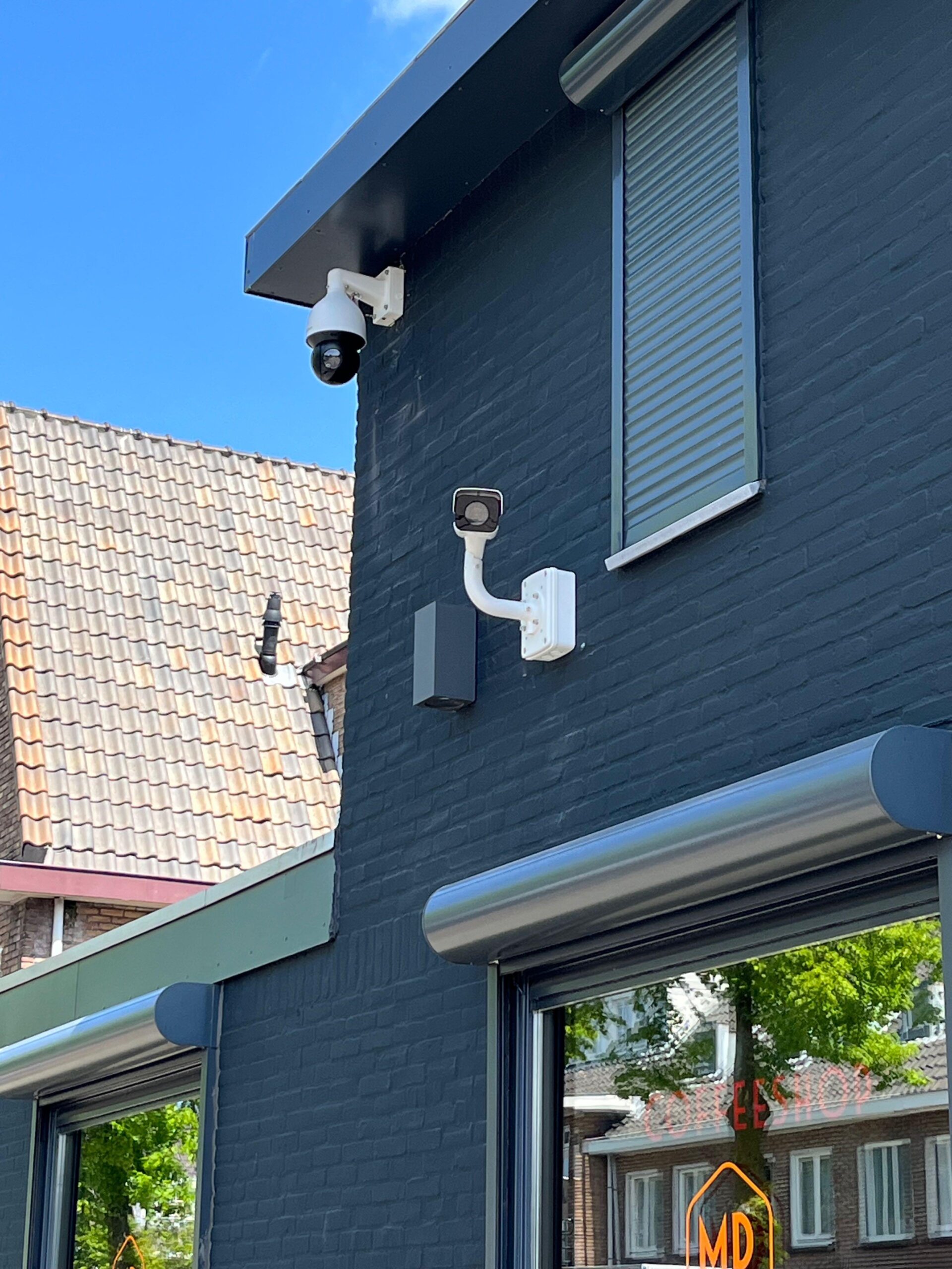 Donkerblauwe gevel van de coffeeshop met bovenaan een witte Dahua PTZ-speeddome onder de dakrand en daaronder een witte bulletcamera op een zwanenhalsbeugel gericht op de straat