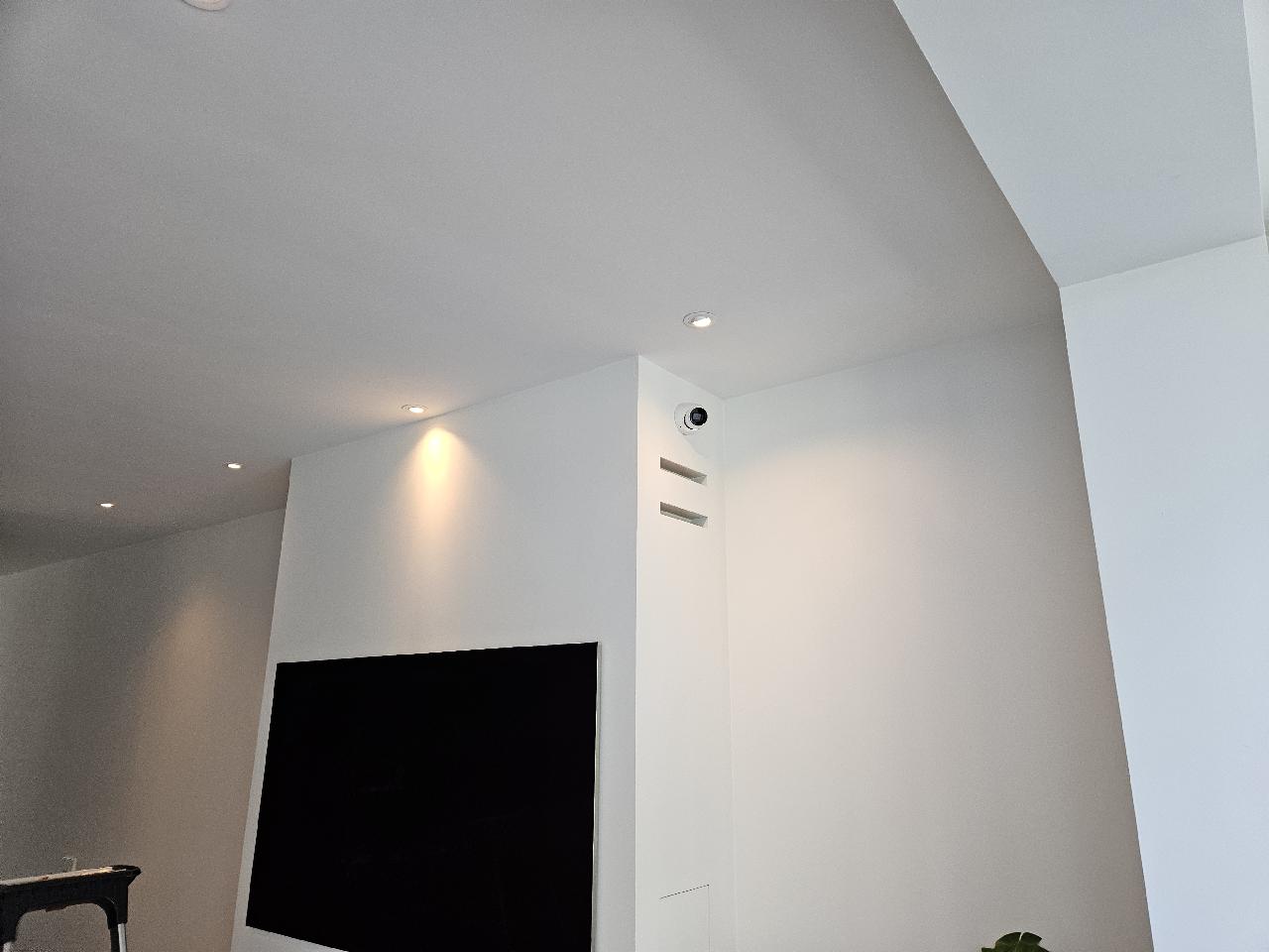 Kleine witte binnencamera hoog in de hoek van een lichte woonkamer, naast een ingebouwd scherm