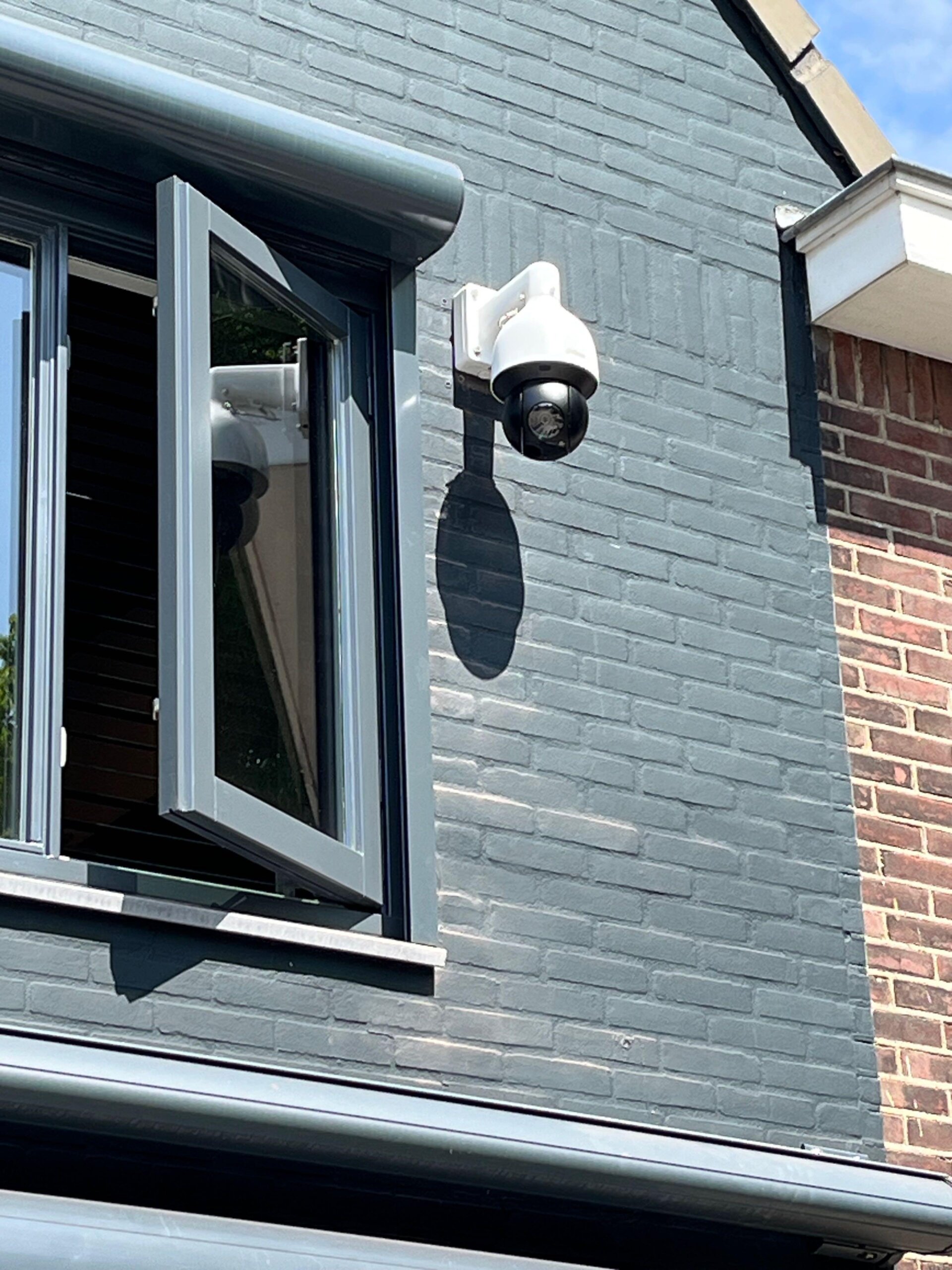 Witte Dahua PTZ-speeddome op een witte wandbeugel, gemonteerd op een grijsblauw geschilderde bakstenen gevel naast een openstaand raam