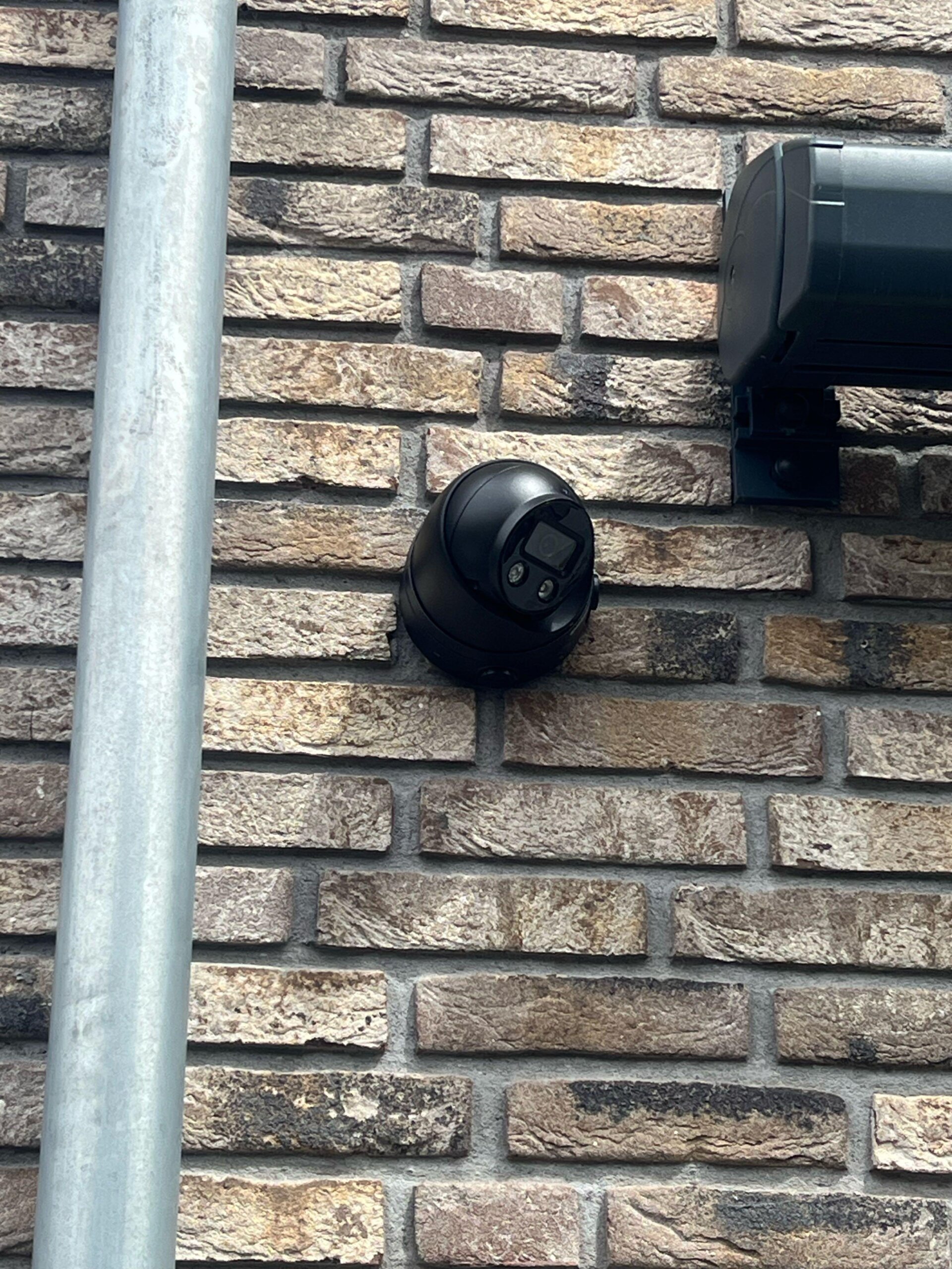 Zwarte camera aan een bakstenen gevel, direct naast een zwarte buitenschijnwerper